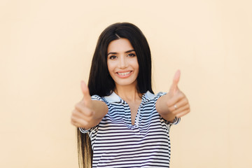 Success and approval concept. Positive female with black long hair, has pleasant smile, keeps...