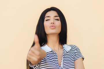 That`s fine! Lovely healthy female with dark hair, looks confidently at camera, raises thumb as shows like gesture, approves something, dressed in casual outfit, isolated over beige background.