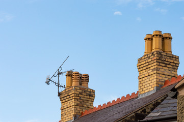 Old residential rooftop and chimneys with TV ariel. Slate roof, decorative red terracotta ridge tiles, yellow brick chimney stacks and chimney pots. Blue sky background with copy space.