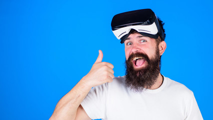 Naklejka premium Man with long beard on happy face showing thumb up. Bearded man with excited look testing new model of VR headset, digital progress concept. Hipster with trendy beard isolated on blue background