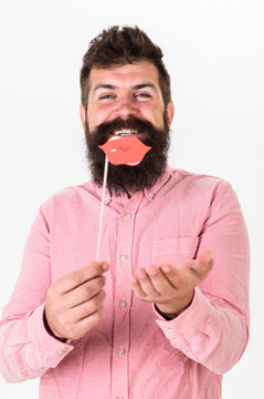 Hipster with beard and mustache on cheerful face posing with photo booth props, copy space. Man holding party props lips, white background. Guy sending air kiss with red lips. Photo booth fun concept