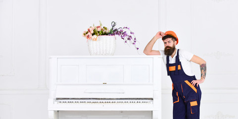 Man with beard, worker in overalls and helmet lean on piano, white background. Courier delivers furniture in case of move out, relocation. Loader moves piano instrument. Delivery service concept.