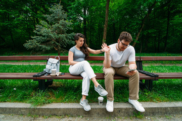 couple sitting on bench in city park. woman is boring, man looking into smartphone.