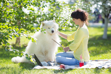 A girl is feeding her dog in a park on a picnic. Beautiful brunette with a big white dog sitting on the grass. The girl gives dog food, and she drinks red juice.