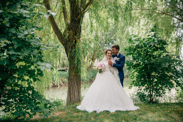 The bride and groom are posing against the background of trees and a pond. A man hugs his beloved...
