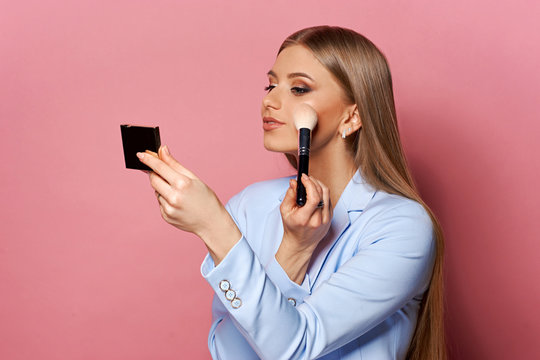 Young Pretty Woman In Blue Suit Holding Makeup Brush And Small Mirror And Applying Cosmetics. Make-up Artist. Girl With Long Straight Blonde Woman Posing Against Pink Background