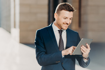 Succesful male entrepreneur in formal suit, holds modern tablet, checks documentation online, reads necessary information for making business report, uses free wifi connection. People and technology