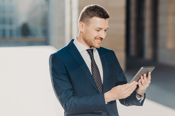 Fototapeta premium Cheerful male manager reads notification on digital tablet, wears formal black suit with tie, connected to wireless internet, watches video on web page. Business and modern technologies concept