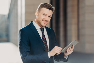 Fototapeta premium Glad confident businessman checks online bank account on touch pad, dressed elegantly, has positive expression, poses outdoors. Executive manager uses modern technologies. People and business