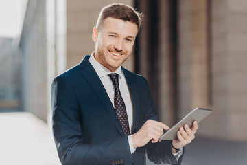 Photo of successful male banker does remote work on digital tablet, has happy expression, dressed in formal black suit, has happy look at camera. Positive young man makes transactions online