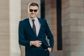 Horizontal shot of handsome cheerful bearded young male manager with confident cheerful expression, keeps hand in pocket, poses against office interior. People, business and elegance concept