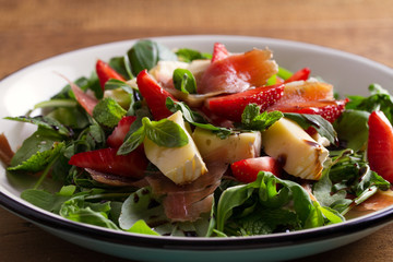 Strawberry, basil, mint, arugula salad with brie cheese and jamon on white plate on wooden table. horizontal