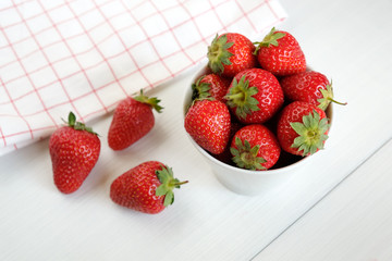 Red ripe strawberries in a cup and checkered dish towel on white wooden table top view.