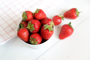Red ripe strawberries in a cup and checkered dish towel on white wooden table top view.
