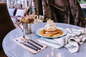 Pancakes with caramel-banana syrup topping with whip cream and mint leaves. With spoon, fork and knife, in blue plate over fabric with blue stripes.