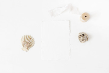 Summer marine, wedding stationery, desktop mock-up scene. Blank greeting cotton paper card, seashells, pebble stone and wooden spool of silk ribbon. White table background. Flat lay, top view.
