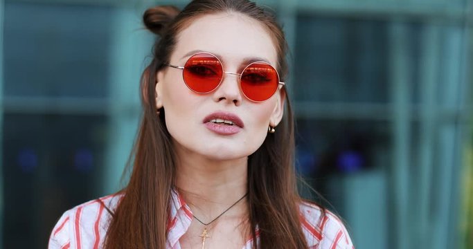 Pretty fashion in red sunglasses poses to the camera with before a modern glass building. A young transgender looking into the camera