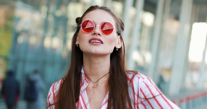 Pretty fashion in red sunglasses poses to the camera with before a modern glass building. A young transgender looking into the camera