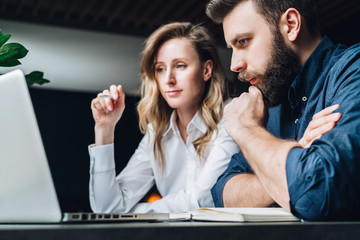 Business meeting. Teamwork. Businesswoman and businessman sitting at table in front of laptop and working. Couple of millennials pondering business project. Online education, e-learning, e-commerce.