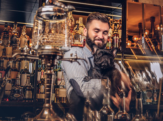 Stylish brutal barman in a shirt and apron keeps thoroughbred black pug at bar counter background.