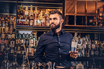 Stylish brutal bartender in a black shirt presents a bottle of exclusive alcohol at bar counter background.
