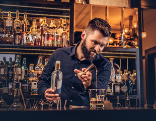 Stylish brutal barman in a black shirt makes a cocktail at bar counter background.