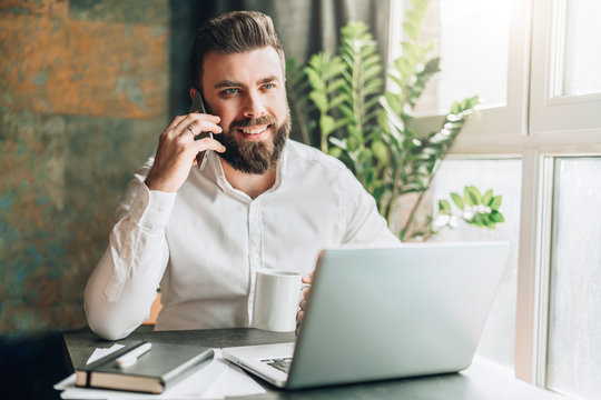 Young smiling bearded businessman is sitting at table in front of laptop, drinking coffee, talking on cell phone. Telephone conversations, distance work, online marketing, education, e-commerce.