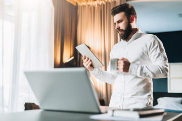 Young bearded bisinessman standing near table in front of laptop, using digital tablet, drinking coffee. Man checks e-mail, blogging, planning. Online marketing, education, e-learning. Social network.