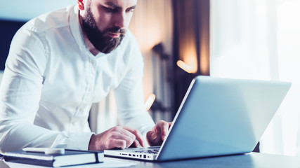 Young serious bearded businessman standing in office near table and using laptop. Man works on computer, checks e-mail, chatting, blogging. Online marketing, education, social network, e-learning.