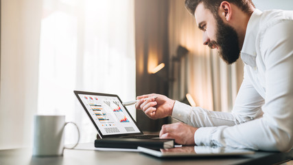Young bearded businessman in white shirt is sitting at table in front of computer, showing pen on laptop screen with graphs, charts, diagrams. Online education, marketing. Business planning.