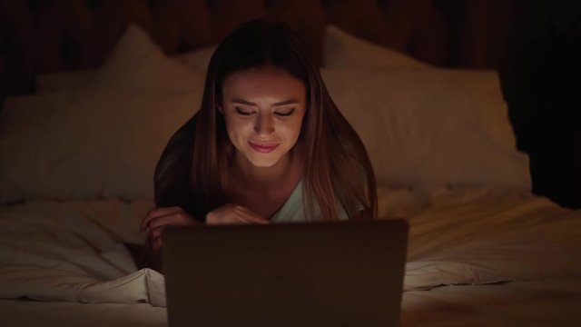 Beautiful young woman with long hair lying on her bed and using her laptop computer at night. Slider real time medium shot
