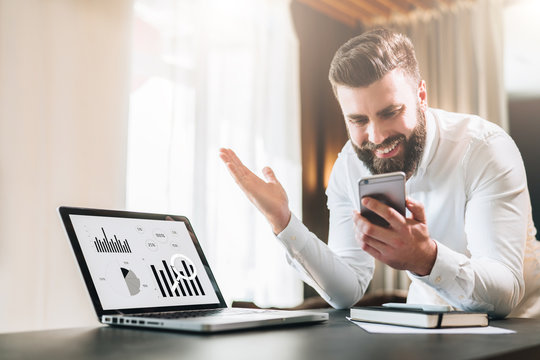 Young bearded businessman in a white shirt is sitting at a table in front of a laptop with graphs, charts, diagrams on screen and is happily looking at screen of smartphone, raising his hands up.