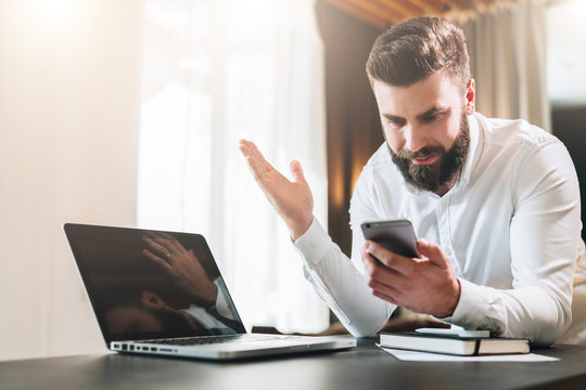 Young bearded businessman in white shirt is sitting at table in front of laptop and is happily looking at screen of smartphone, raising his hands up.Online marketing, education, e-learning,e-commerce.