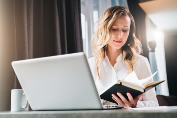 Young businesswoman in shirt is sits in office at table in front of computer and reads notes in notebook.Student does homework, girl reads book. Online marketing, education, e-learning, distance work.