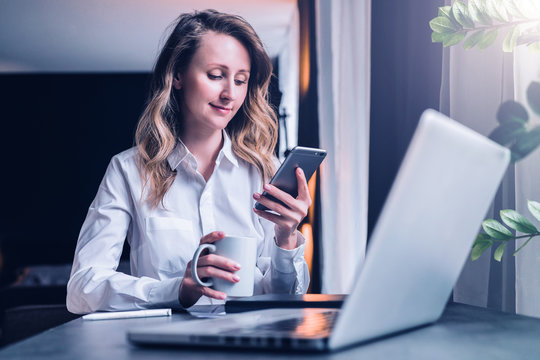 Young businesswoman in shirt is sitting in office at table in front of computer, using smartphone, looks at phone screen, holds cup of coffee in her hand. Social network, breaking. Student learning.