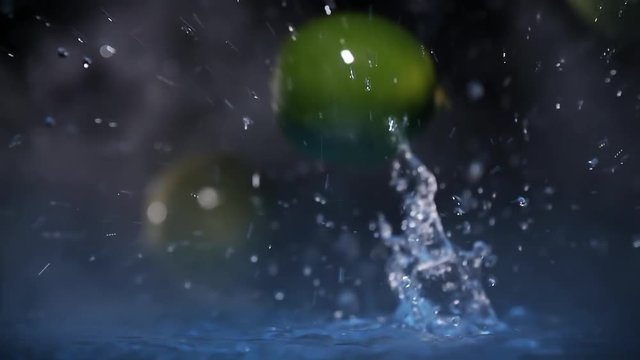 Lime fruits falling on water surface in blue light spot and cold fog cloud with splash and drops close up