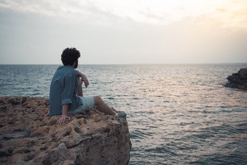 Charming dreamer. Full length back view of pensive stylish bearded male is sitting on the edge of cliff with endless ocean in background. He is looking at water thoughtfully. Copy space in right side