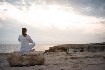 Fototapeta na wymiar Inner harmony. Full length back view of calm girl is sitting on beach while meditating. She is practicing yoga on beach while enjoying wonderful view on the sea. Copy space in the right side