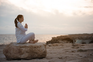 Fototapeta premium Harmony and relaxation. Full length side view profile of young woman in white clothes is sitting on big stone. She is keeping legs crossed while practicing asana with closed eyes. Copy space