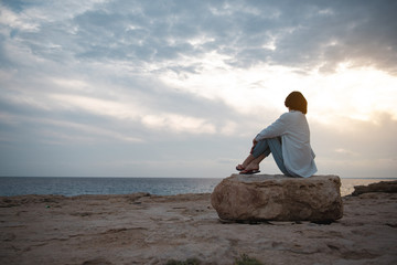 Obraz premium Cudowny widok. Pełny widok z tyłu przemyślanej dziewczyny na plaży siedzi na dużym okrągłym kamieniu, ciesząc się zachodem słońca. Patrzy na ocean i myśli sennie. Skopiuj miejsce po lewej stronie