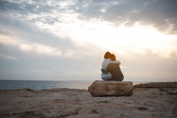 Full length back view of two pleasant women are sitting on the beach on big stone and having strong hug. They are looking at ocean while enjoying sundown. Copy space in the left side