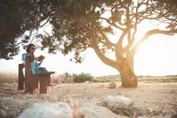 Full length of young attractive dreamy woman in sunglasses is sitting on bench and reading book with concentration. She is resting near big old tree against wonderful sunset. Copy space in right side