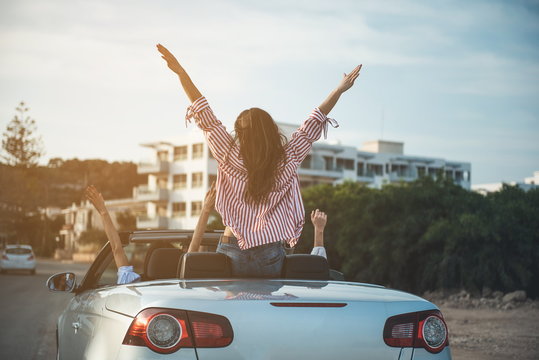 Freedom. Back view of happy positive friends are travelling in fashionable cabriolet with open roof. They are sitting in car with raised hands while enjoying summer vacation. Luxury lifestyle concept
