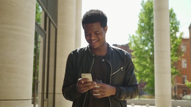 Handsome African American man in leather jacket standing outdoors in sunlight and enjoying texting on his phone, coffee cup in his hand