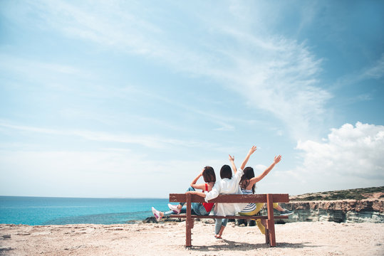 Fun together. Full length back view of three cheerful girls are sitting on bench and looking at azure ocean. They are expressing gladness with raised hands. Summer trip concept and copy space