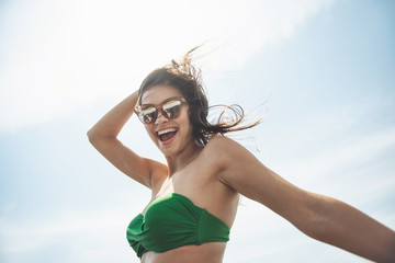 Happy day. Low angle portrait of young cheerful attractive woman in sunglasses and swimsuit is standing outdoors and enjoying summer vacations abroad. She is looking at camera with joy. Travel concept