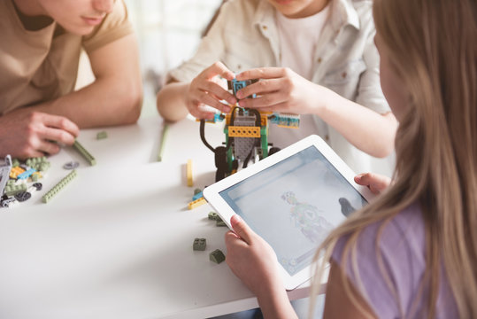 Girl looking at screen of electronic tablet. Child making toy near her. They sitting at table