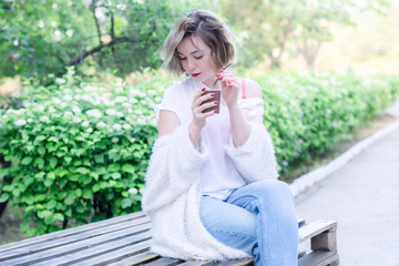 Attractive girl with red lips is sitting in the park with paper cup of coffee