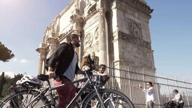 Three young friends tourists walking with bikes near constantine arch at Colosseum in Rome city centre on sunny day slow motion camera steadycam ground shot