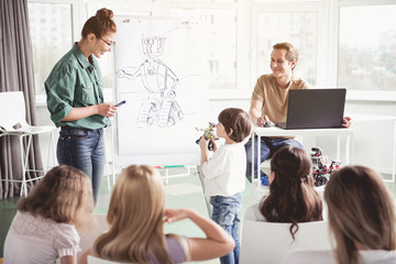 Positive boy answering happy woman while showing at robot. He keeping it in hands. Cheerful man using gadget during lesson. Happy lesson concept